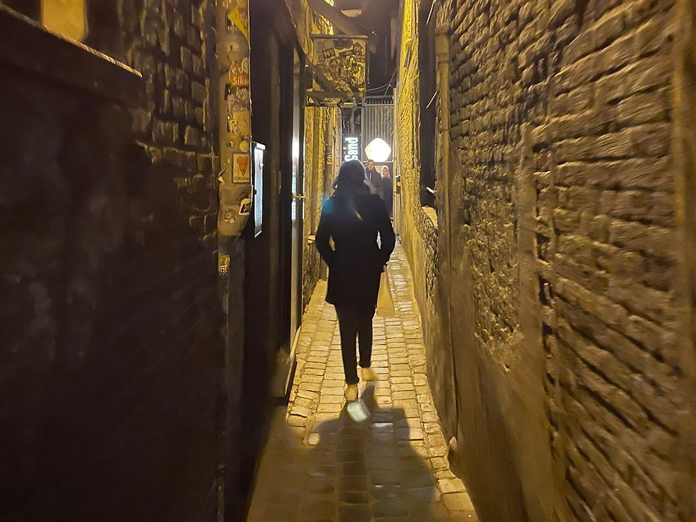Person walks down a narrow, dimly lit cobblestone alley flanked by brick walls adorned with stickers. Warm yellow light creates a moody atmosphere.