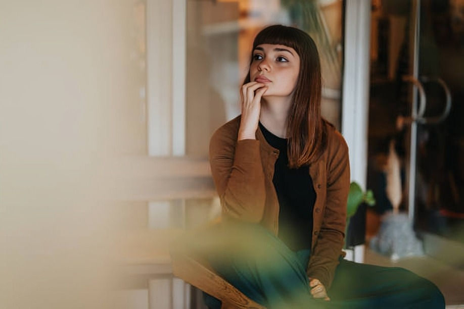 Thoughtful young woman in a brown cardigan sits indoors, hand on chin, with a soft blurred foreground and warm light.