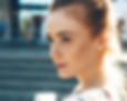 A young woman with freckles and red hair tied back gazes thoughtfully to the side, bathed in warm sunlight with a blurred urban background.