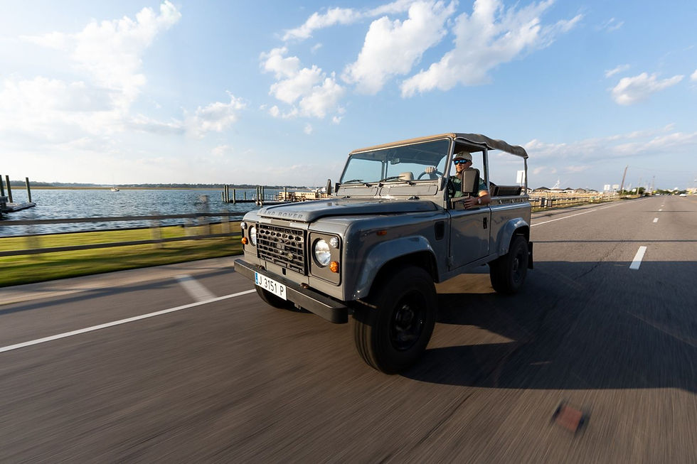 Convertible Land Rover drives on a coastal road, driver wearing sunglasses. Blue sky with clouds and water in the background, sunny.