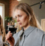 Woman in a gray shirt smelling red wine in a glass, appearing focused. Indoors with a cozy background featuring plants and warm lighting.