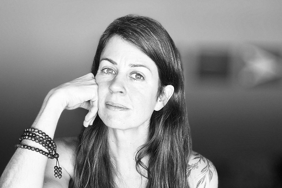 Woman with long hair and tattoos leans on hand, wearing beaded bracelets. Black and white image, neutral expression, blurred background.