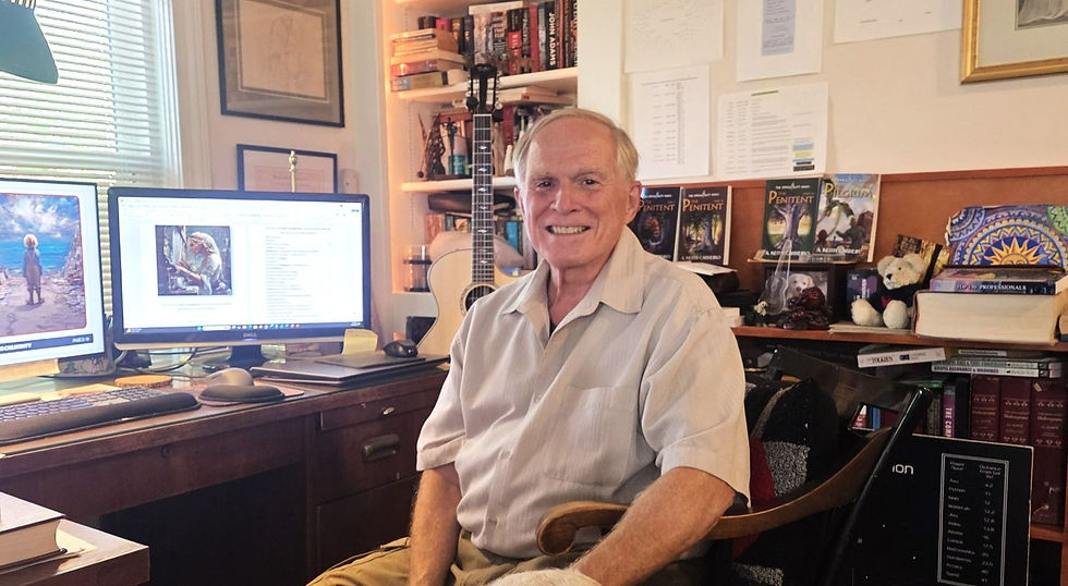 Smiling older man sits in a cluttered home office with dual monitors, books, papers, and a guitar behind him.