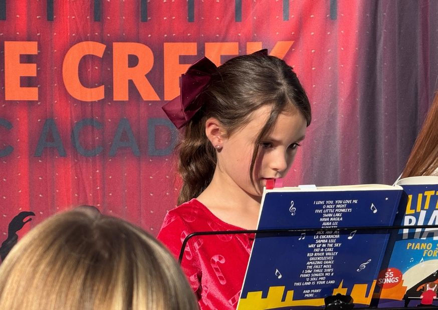 Young girl in red dress with bow reads piano book on stand. Red curtain backdrop with "CREEK" visible. Focused and calm mood.