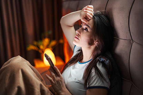 Woman in bed with white shirt holds smartphone, looking stressed. Background has warm lamp and curtains, creating a cozy evening setting.
