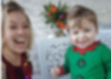 Smiling woman in red with festive lights and cheerful toddler in green outfit pose indoors. White brick wall and colorful flowers behind.