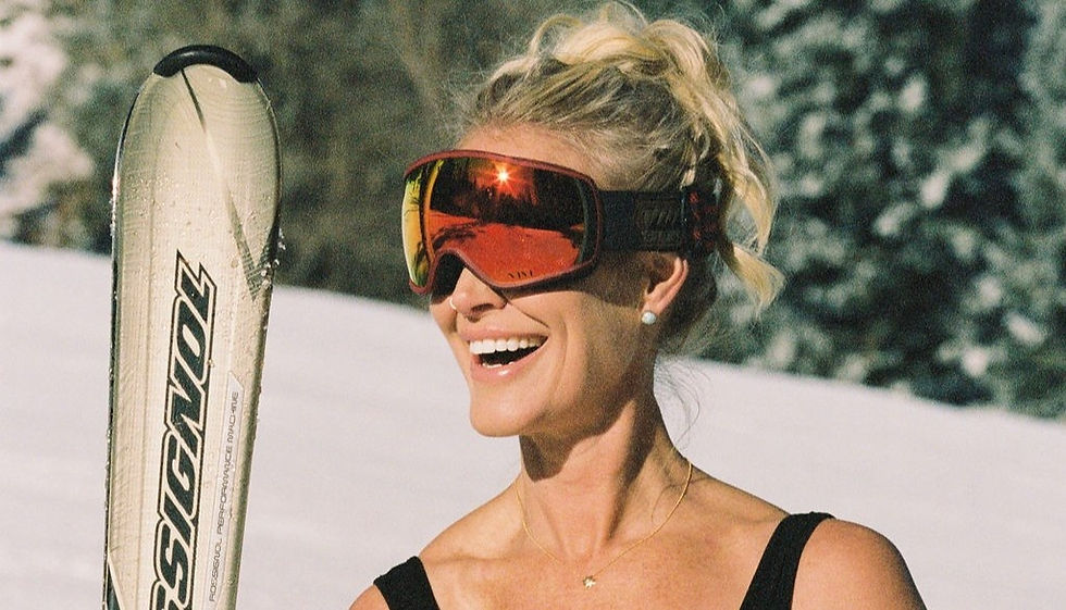 Smiling blonde woman in ski goggles holds a Rossignol ski on a snowy slope, with dark trees behind.