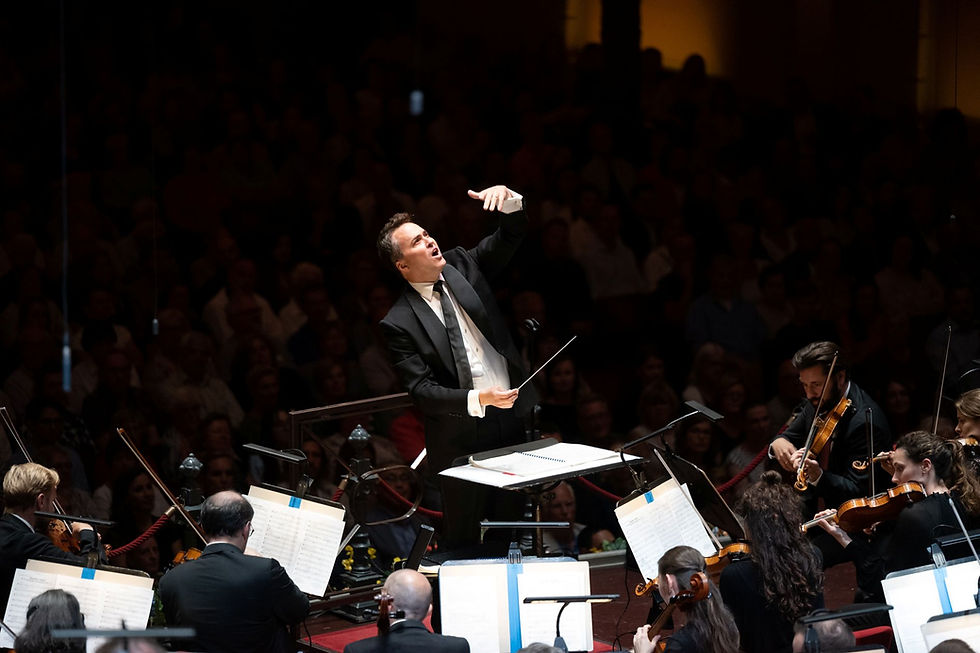 Garrett Keast passionately leads an orchestra, surrounded by musicians with violins. The dimly lit concert hall is filled with an attentive audience.