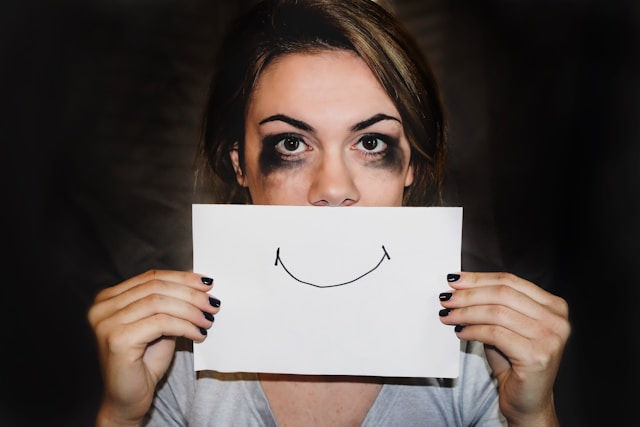 Woman with smudged makeup holds a drawn smile on paper over her mouth. Dark background, conveying contrast between emotion and appearance.