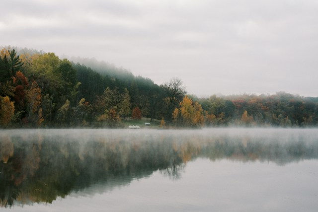Foggy lake reflecting autumn trees and cloudy sky, with mist drifting low over calm water and a quiet, peaceful mood.