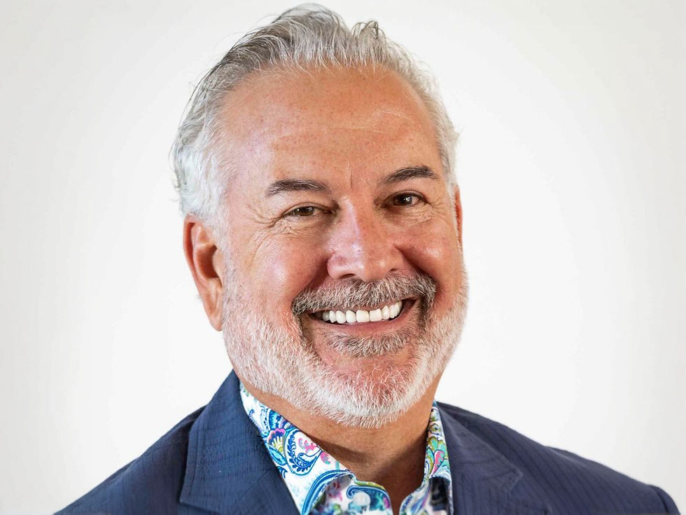 Smiling man with gray hair and beard in a blue suit, colorful paisley-patterned shirt, against a plain white background.
