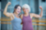 Two women in workout gear smile and flex their arms in a gym setting, showcasing strength and happiness. Blurred background.
