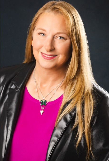 Smiling blonde woman in a black leather jacket and hot pink top, wearing a butterfly necklace, against a dark studio background.