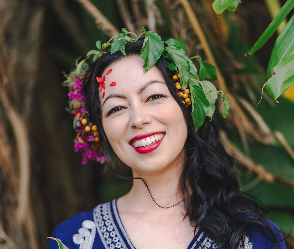 Smiling woman with leaf and berry headpiece, vibrant red lipstick, and embroidered blue dress, set against lush green foliage.