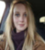 Woman with long blonde hair in a car, wearing a brown coat and purple scarf. She has a calm expression, with soft lighting around.