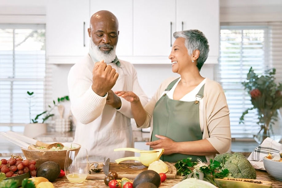 Smiling older couple cooking together in a bright kitchen, surrounded by fresh produce and bowls on the counter.