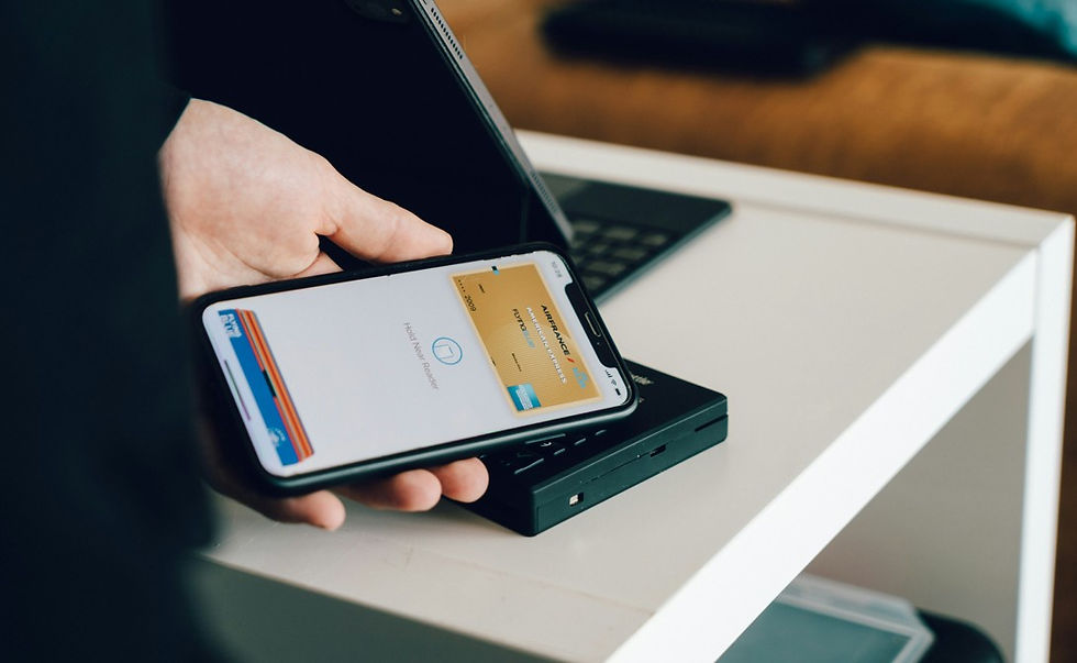Hand holding smartphone over a card reader on a white table, with a laptop in the background; payment app screen visible.
