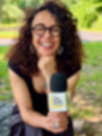 Curly-haired woman smiling, holding a microphone labeled "The Lulu Essey Podcast" in a sunny park. Bright green foliage in the background.