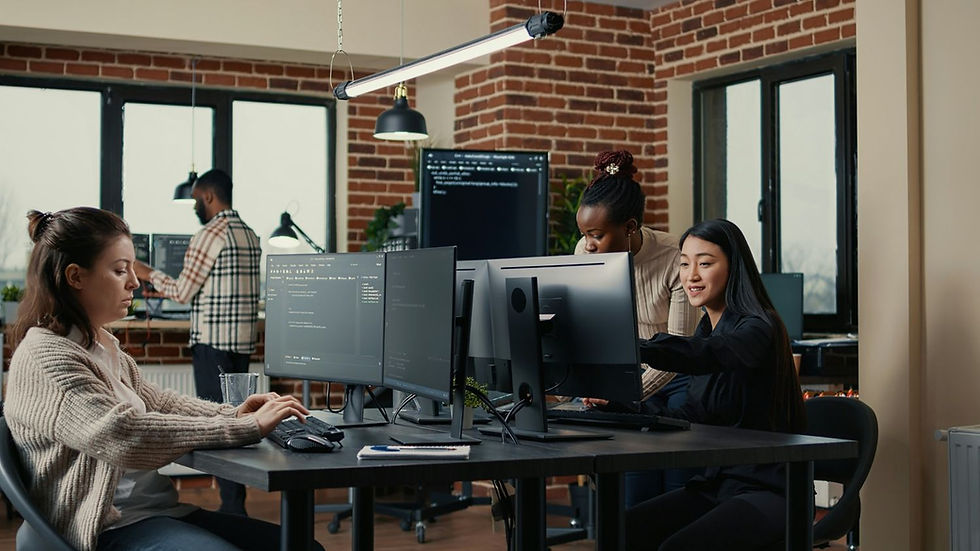 Three people working on computers in an office with brick walls. One woman types, another points at a screen, a man stands in the background.