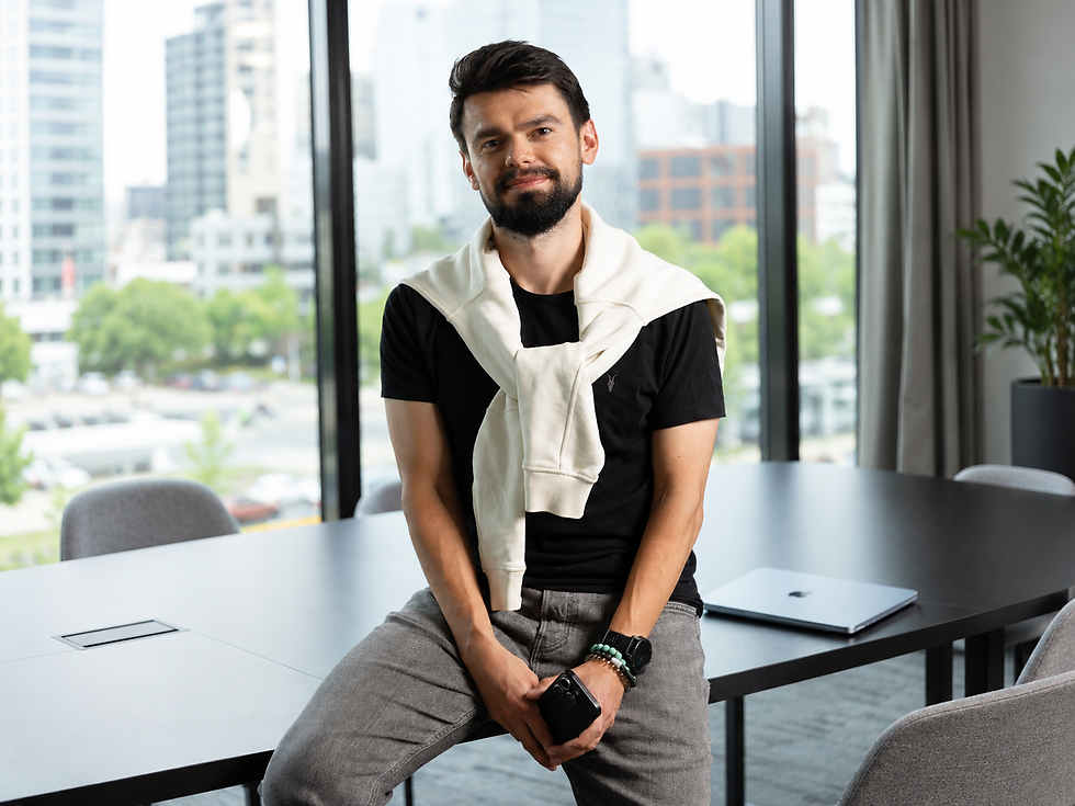 Smiling man sits on a conference table in a modern office, holding a phone; laptop nearby, city skyline behind him.
