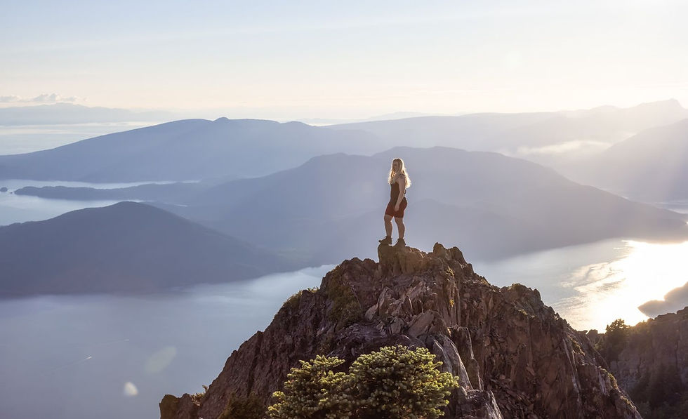 Woman stands on rocky mountain summit, overlooking expansive view of misty blue mountains and shimmering water at sunrise. Calm and serene.