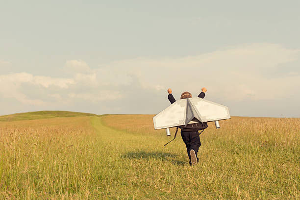 A young business boy is wearing a jetpack and running through a grass field.