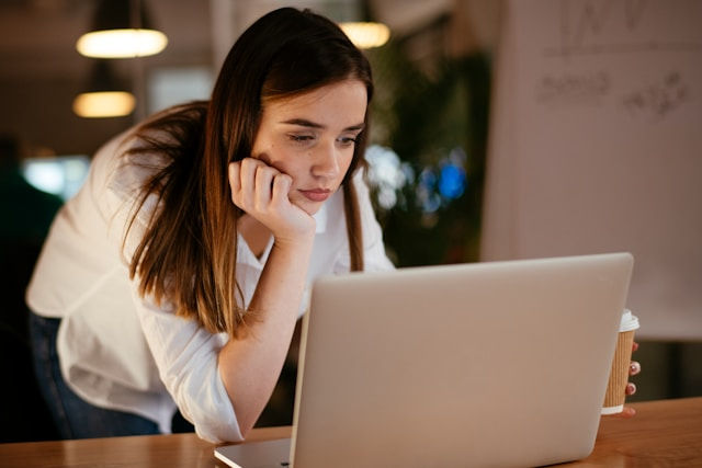 Woman in white shirt leans on a table, looking at a laptop screen, holding a coffee cup. Soft lighting creates a focused atmosphere.