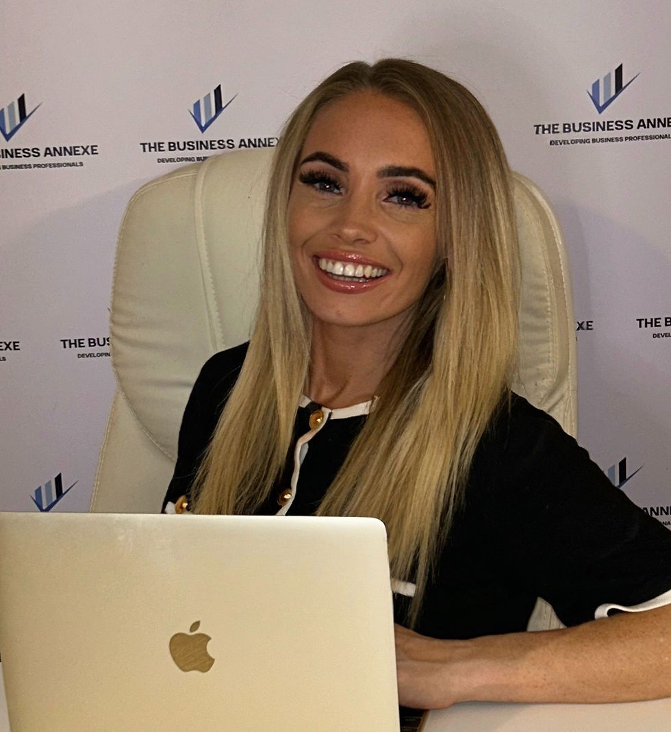 Smiling woman with long blonde hair sits at a desk with an Apple laptop. Background shows "The Business Annexe" logo on the wall.