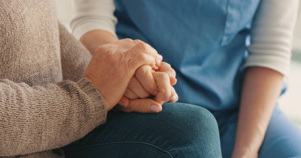 Elderly person holding a caregiver’s hand in a comforting close-up, with blue scrubs in the background.