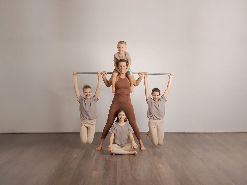 Five kids pose in a studio. Two hang from a bar, one sits on a shoulder, and two stand or kneel. Neutral colors, wooden floor.