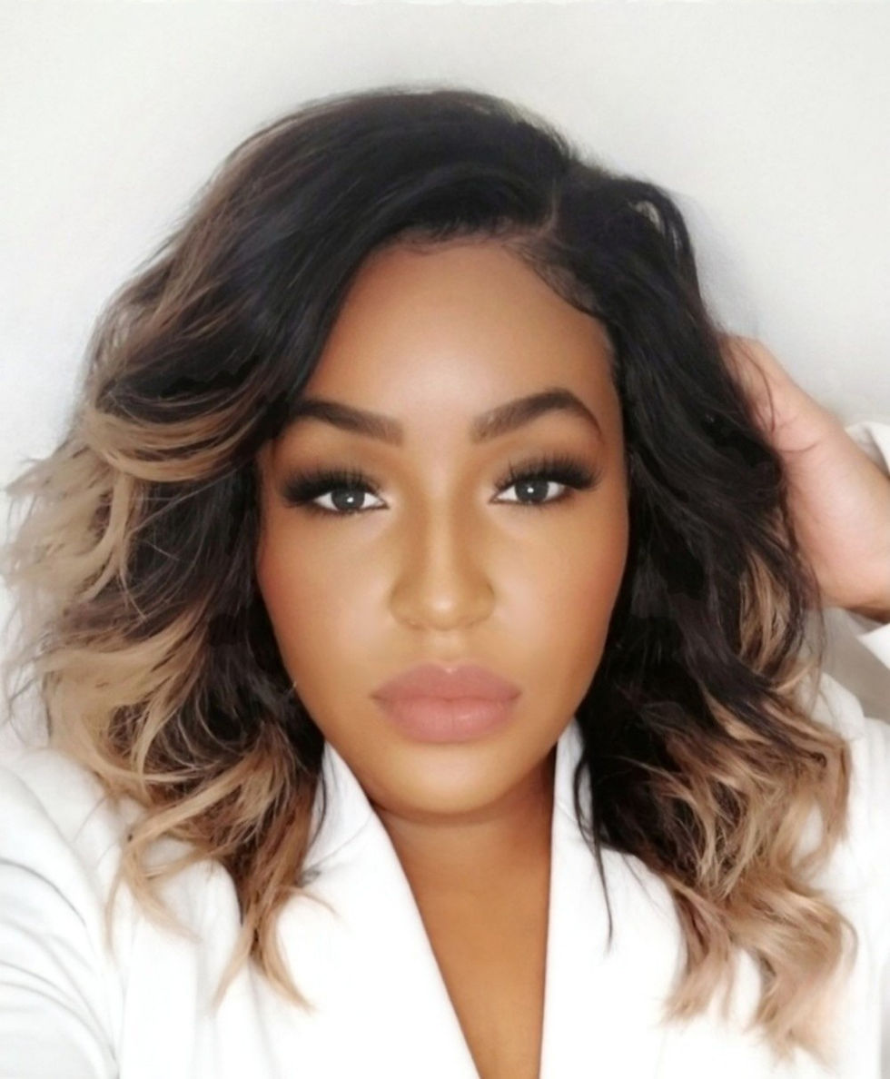 Woman with wavy black and blonde hair poses against a plain background, wearing a white top. She has a neutral expression.