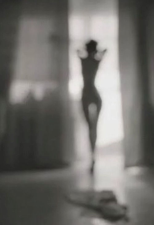 Silhouette of a person stretching against a bright window, with clothes on the floor. Blurred grayscale image creates a serene mood.