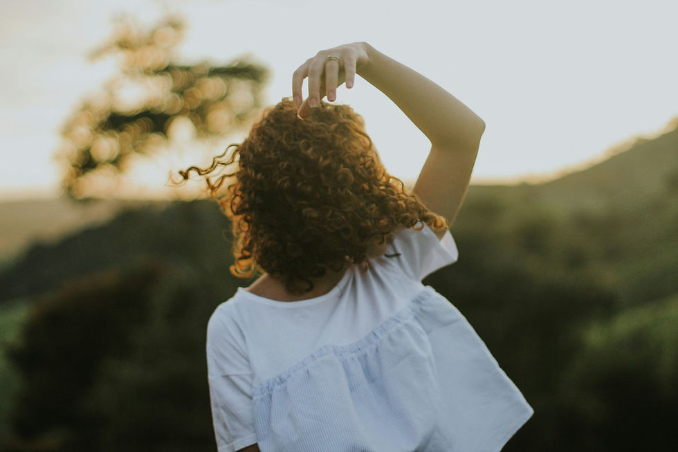 Curly-haired person in white shirt raises arm, backlit by sunset. Blurred trees and hills create a serene, golden atmosphere.