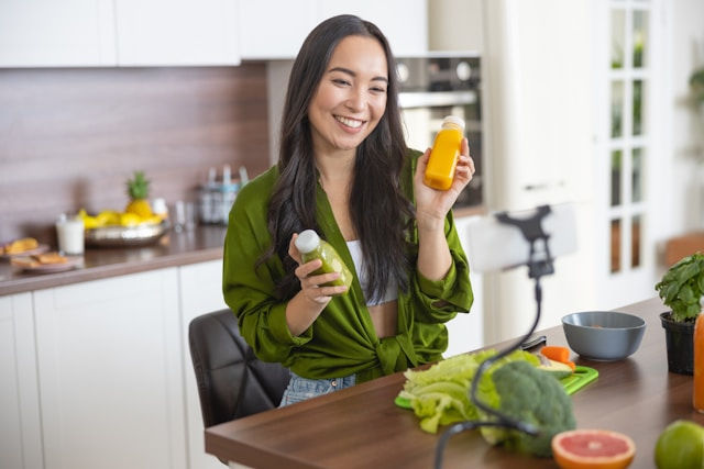 Smiling woman in a kitchen holds green and orange juice bottles while filming with a phone. Fresh veggies and fruits are on the table.