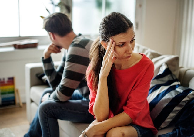 Tense couple sits apart on a sofa, woman in red holding her head, man in striped shirt turned away in a bright living room.