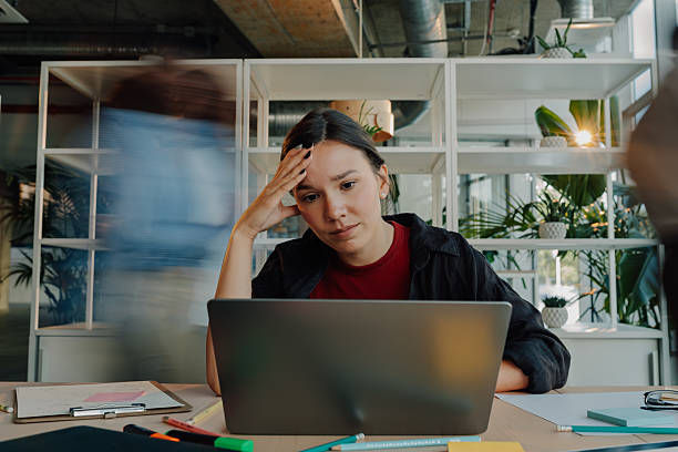 Tired woman at a laptop in an office, resting her head while a blurred coworker passes behind her and plants fill shelves.