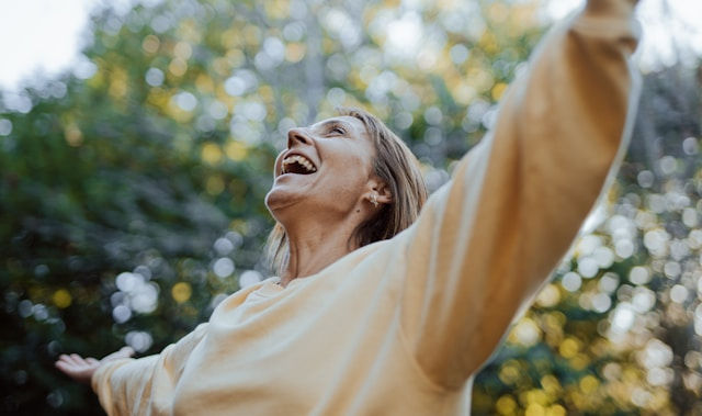 Smiling woman in a pale sweater raises her arms outdoors, laughing against a sunlit, blurry tree background.