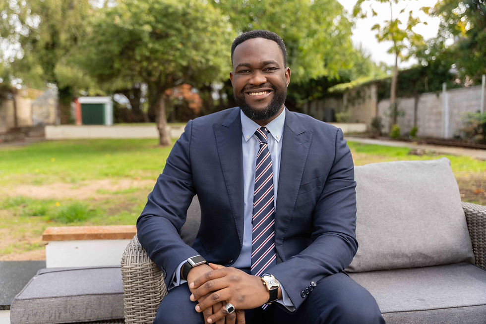 Smiling man in a navy suit and striped tie sits on a gray sofa in a lush garden, conveying a cheerful, relaxed mood.