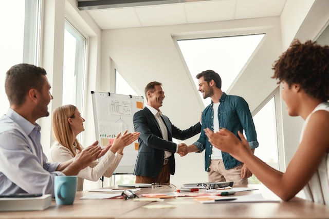 Five people in an office, two men shaking hands, others clapping. A flipchart with graphs is in the background, sunlight through windows.