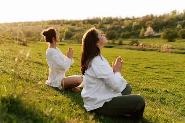 Two women are meditating outdoors in a grassy field at sunset, sitting peacefully with their hands in prayer position.