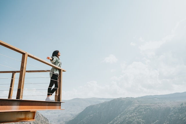 Woman with outstretched arms stands on a wooden balcony, overlooking a vast mountain landscape under a clear, bright sky. Feeling of freedom.