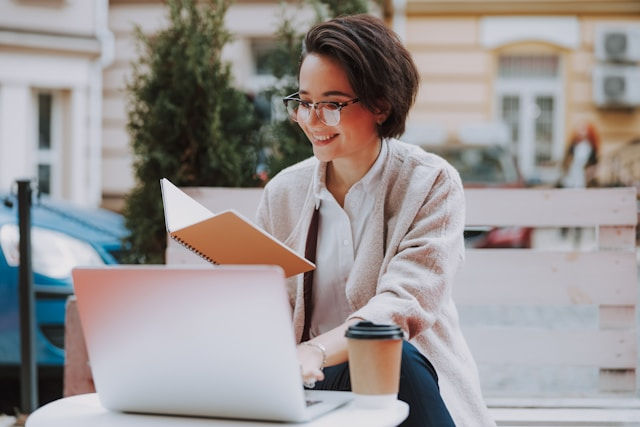 Smiling person with glasses holds a notebook, sitting at an outdoor table with a laptop and coffee. Background shows blurred buildings.