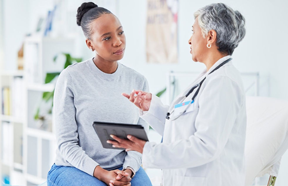 Doctor in white coat talks with seated patient in a bright clinic, holding a tablet; calm, attentive consultation.