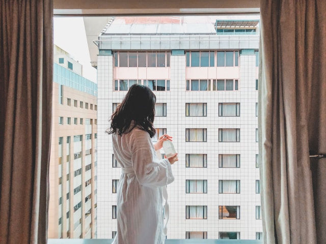 Woman in a white robe holds a drink, stands by a window with city building view. Curtains frame the scene, creating a calm, reflective mood.