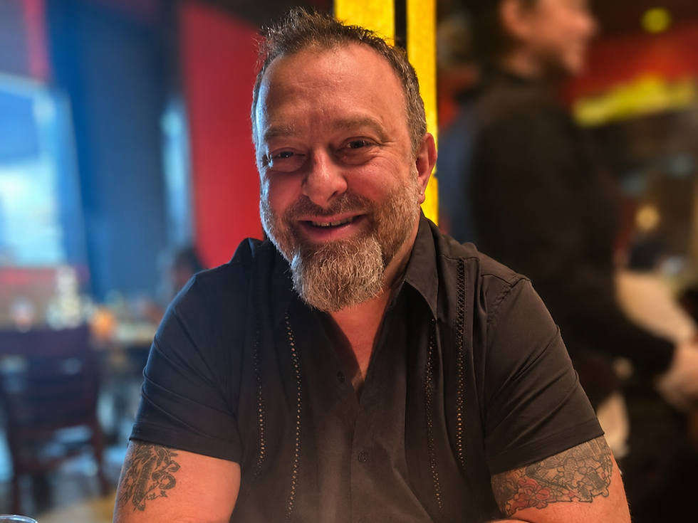 Smiling tattooed man in a black shirt sits in a dim, colorful bar or restaurant.