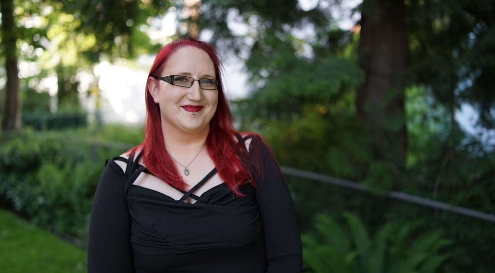 Smiling person with red hair and glasses in a black top, standing outdoors with lush greenery in the background.