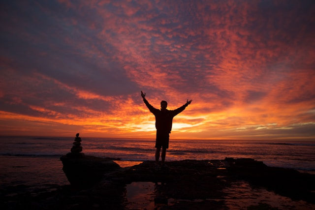 Silhouette of a person on rocks with arms raised at a vivid orange-purple sunset over the ocean, with stacked stones nearby