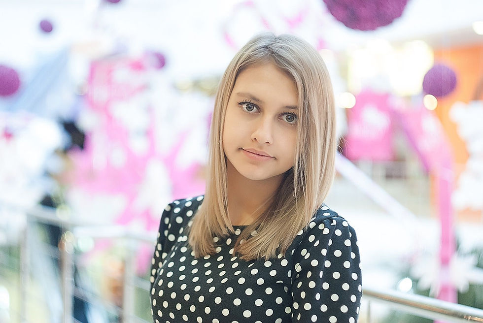 a young woman with straight blonde hair, wearing a black polka-dotted shirt, standing in an indoor setting with soft, colorful decorations in the background.