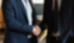 Two businessmen in dark suits shake hands in a modern office, suggesting a formal agreement.