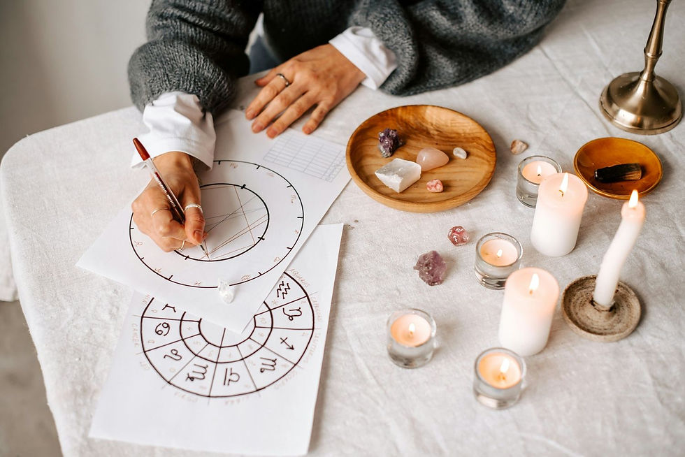 Person drawing on an astrology chart with candles, crystals, and wooden dish on a white tablecloth. Calm and focused atmosphere.